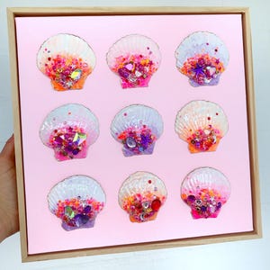 May include: A square wooden frame containing a light pink background with nine white scallop shells arranged in a grid. Each shell is decorated with colourful beads, sequins, and faceted crystals in shades of pink, purple, orange, and red.