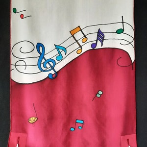 May include: A white and red prayer shawl with a colorful musical note design. The shawl has tassels on the bottom corners.