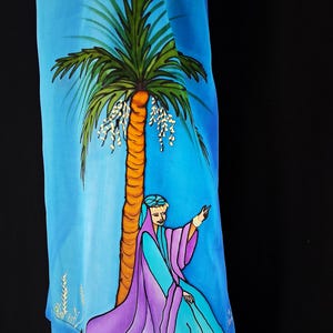Peut inclure: Un tissu bleu avec une scène peinte représentant un palmier et une figure en robes violettes et turquoise. Le palmier a un tronc brun et des frondes vertes. La figure est agenouillée sous l'arbre, un bras levé.