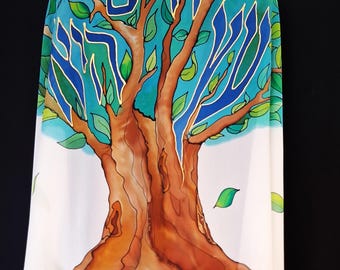 May include: A white shawl featuring a hand-painted tree with a brown trunk and green leaves against a blue sky background. The Hebrew text "חיים" and "שלום" is painted in blue with gold outlines integrated into the branches of the tree.