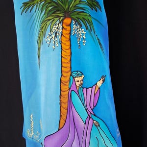 May include: A blue silk scarf featuring a hand-painted illustration of a palm tree and a figure wearing a purple and turquoise robe. The figure is seated at the base of the tree, pointing upward with one hand, set against a bright blue background.