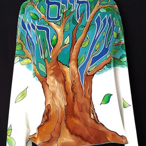 May include: A white shawl featuring a colorful tree design. The tree has a brown trunk, blue and green leaves, and blue Hebrew text at the top. The shawl is draped over a black garment.