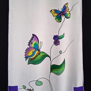 May include: A white textile featuring a hand-painted design of two colorful butterflies with intricate wing patterns. The butterflies are set against a backdrop of green leaves, purple flowers, and delicate black vines. The artwork is signed in the lower corner.
