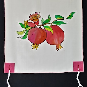 May include: A white fabric with a hand-painted design of pomegranates. The pomegranates are red with green leaves and yellow flowers. The corners have pink squares with white strings attached.