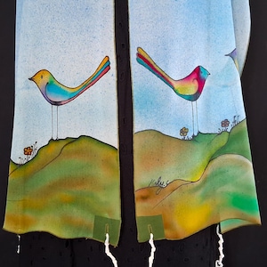 May include: A colorful, hand-painted tallit with a whimsical design. The artwork features vibrant birds perched on rolling hills with flowers, set against a light blue sky. The tallit has a black background.
