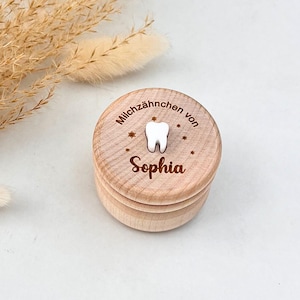 Könnte beinhalten: Kleine, runde Andenkenbox aus hellem Holz für Milchzähne. Der Deckel ist mit einem Zahnmotiv, Sternen und dem Namen "Sophia" graviert. Der Text "Milchzahnchen von" ist ebenfalls eingraviert. Ein schönes Geschenk.