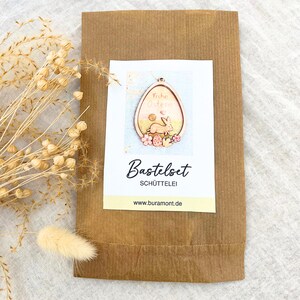 May include: A brown paper bag with a white label featuring an Easter egg design. The egg-shaped design includes a bunny, flowers, and the words "Frohe Ostern." The label also says "Bastelset SCH&Uuml;TTELEI."