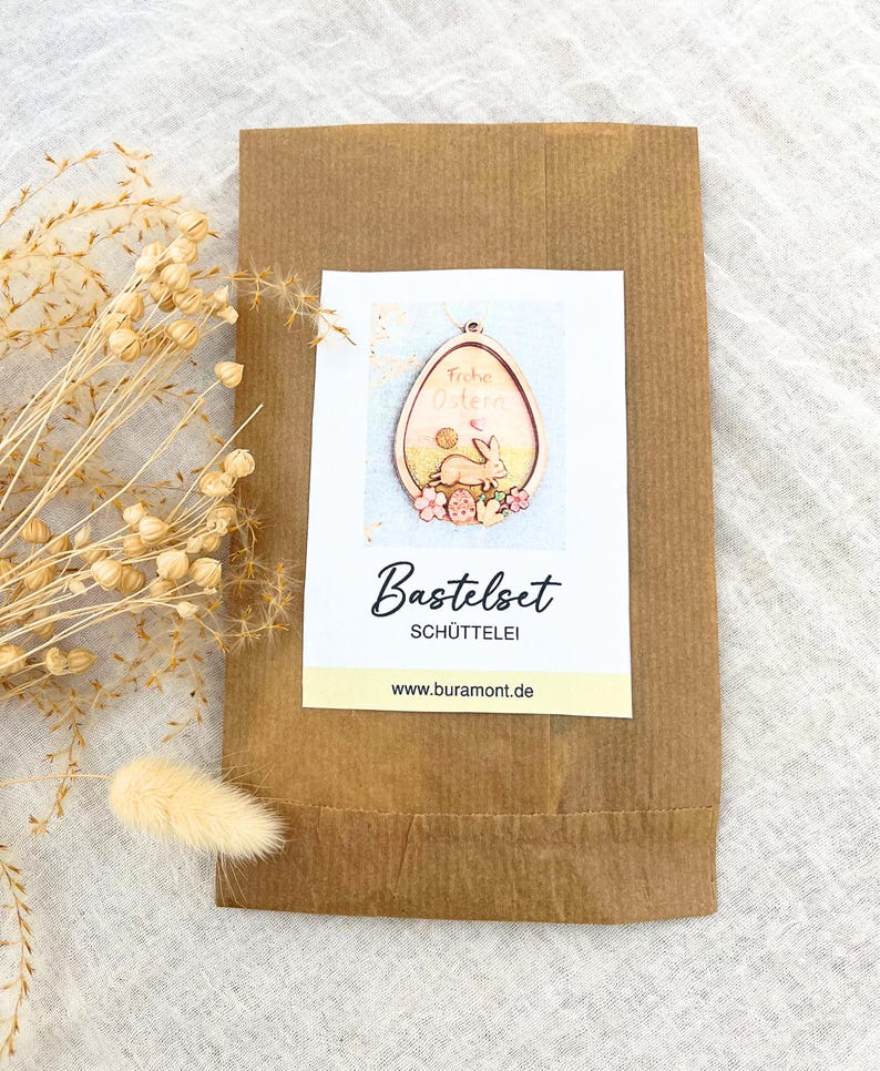 May include: A brown paper bag with a white label featuring an Easter egg design. The egg-shaped design includes a bunny, flowers, and the words "Frohe Ostern." The label also says "Bastelset SCH&Uuml;TTELEI."