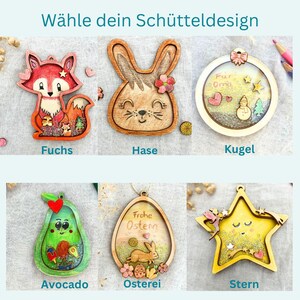 May include: A collection of wooden ornaments in various shapes, including a fox, bunny, avocado, and star. Each ornament features intricate details and vibrant colors, with some containing small decorative elements. The text "W&auml;hle dein Sch&uuml;tteldesign" is at the top.