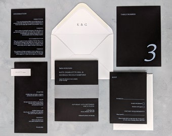 May include: Black and white wedding invitation suite with a silver accent. The invitation includes the names "Kate Charlotte Hill & Georgia Nicole Bamford", the date "Saturday 24th September", and the time "One O'Clock". The RSVP card includes the text "RSVP", "Name(s)", "Dietary Requirements", and a space for guests to indicate their acceptance or decline. The table number card is black with a white number "3".