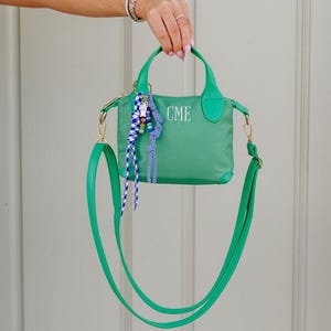 May include: A green handbag with a detachable shoulder strap and top handles. The bag features the embroidered initials "CME" and is adorned with a decorative tassel. The bag is held up by a person's hand.