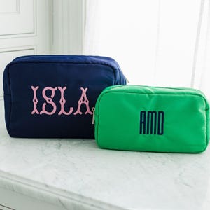 May include: A navy blue rectangular pouch with the name ISLA embroidered in light pink thread and a smaller green rectangular pouch with the initials AMD embroidered in dark navy blue thread, both featuring top zipper closures.