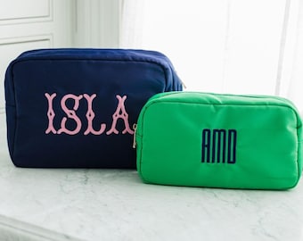 May include: A navy blue rectangular pouch with the name ISLA embroidered in pink and a smaller green rectangular pouch with the initials AMD embroidered in navy blue. Both pouches feature a zipper closure and a structured fabric design.
