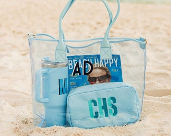 May include: A clear tote bag with light blue accents, filled with beach essentials. Includes a light blue tumbler, a magazine with the text "BEACH HAPPY", and a light blue cosmetic bag with "CHS" embroidered on it. The items are on a sandy beach.