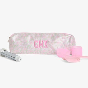 May include: A cosmetic bag with a pink floral pattern and the embroidered initials "CME" is displayed with a curling iron, pink hair rollers, and a pink hairbrush. The bag has a zipper closure.