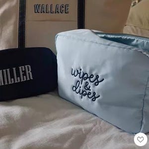May include: A collection of personalized bags and pouches. A large beige tote bag with black handles and the name "WALLACE" embroidered in black. A navy blue pouch with "MILLER" and a light blue pouch with "wipes & dipes".