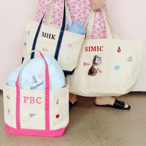 Op de afbeelding: Drie canvas draagtassen in off-white met gekleurde randen en hengsels. Eén heeft marineblauwe randen en 'MHK', één heeft roze randen en 'PBC' en één heeft off-white randen en 'SIMIC'. Elke tas is voorzien van geborduurde patches met diverse iconen.