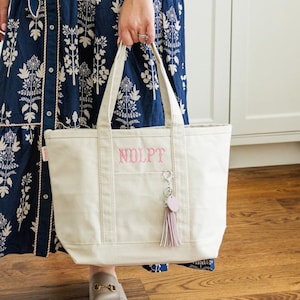 May include: A navy blue dress with a white floral pattern and a matching belt. The dress has short sleeves and a button-down front. A cream-colored tote bag with the letters "NOLPT" is held in front of the dress. The bag has a pink tassel.