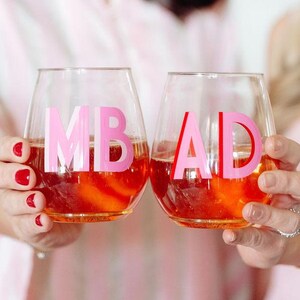 Könnte beinhalten: Zwei Weingläser ohne Stiel, gefüllt mit einer rot-orangen Flüssigkeit. Jedes Glas hat rosa Buchstaben: "MB" und "AD". Die Gläser werden von zwei Personen hochgehalten, was auf ein festliches Ereignis hindeutet. Das Gesamtthema bezieht sich auf Partys und Zusammenkünfte.