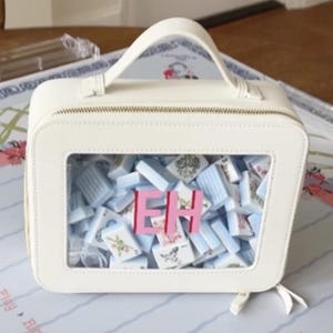 May include: A cream-coloured rectangular handbag with a clear front panel, filled with blue and white tiles and pink letters spelling "EH". The bag has a top handle and a zip closure. It rests on a floral patterned surface.