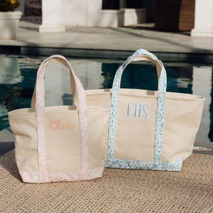 May include: Two canvas tote bags with handles. One has pink floral trim and the name "Olivia" embroidered in orange. The other has blue floral trim and the initials "CHS" embroidered in blue. Both bags are beige.