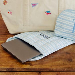 May include: A cream-colored canvas tote bag with the name CONDER printed in black, featuring various small embroidered patches. Beside it is a light blue and white wavy-striped zippered pouch with the monogram TB and a matching laptop sleeve.