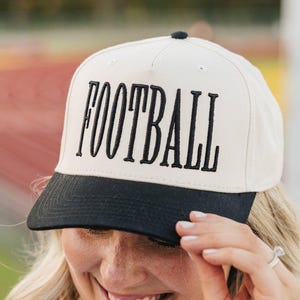 Puede incluir: Gorra de béisbol color crema con visera negra y la palabra "FOOTBALL" bordada en letras mayúsculas negras. La gorra tiene un botón negro en la parte superior.