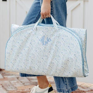 May include: A floral garment bag with a light blue trim and handles. The bag features a light blue monogram and a delicate floral pattern on a white background. The person is wearing blue jeans and white sneakers.