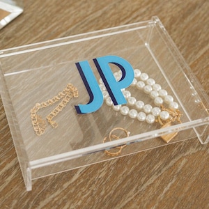 May include: A clear acrylic jewelry box with a blue and white monogram "JP" on the lid. The box contains a pearl necklace, a gold chain, and a small gold ring.