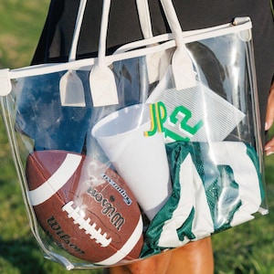 May include: A clear tote bag with white trim and handles, filled with sports gear. Inside are a brown football, a white cup, and a green and white jersey. The bag is held by a person wearing a black dress.