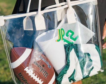 May include: A clear tote bag with white trim and handles, filled with sports gear. Inside are a brown football, a white cup, and a green and white jersey. The bag is held by a person wearing a black dress.