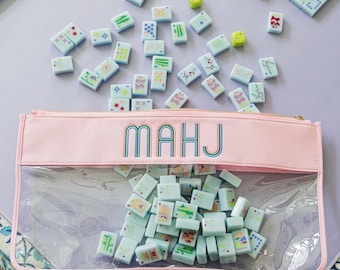 May include: A Mahjong set with light blue tiles scattered on a lavender surface. A pink pouch with "MAHJ" printed on it is filled with tiles. The table has a floral patterned border and matching game rails.