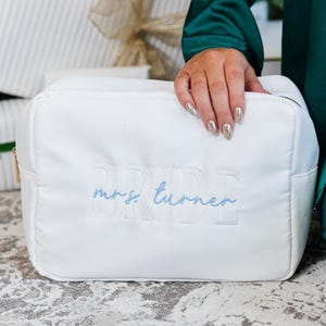 May include: A white rectangular cosmetic bag featuring the word BRIDE embroidered in white block letters and the name mrs. turner embroidered in light blue cursive script across the center of the front panel with a gold zipper closure.