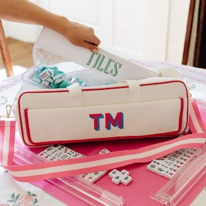 May include: A white rectangular mahjong bag with pink trim and a pink and blue monogram TM on the front. The bag has a pink and white striped shoulder strap and contains a clear pouch labelled TILES. Several white mahjong tiles are visible.