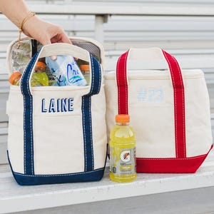 May include: Two canvas cooler bags, one with "LAINE" embroidered in blue, the other with "#23". The bags are off-white with blue and red accents. A bottle of yellow sports drink sits nearby. The bags are on a metal bench.