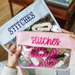 May include: Two clear-windowed pouches, one light blue and one pink, each with the word "stitches" embroidered on the front. The pouches contain embroidery floss and other craft supplies. Gold zippers secure the pouches.