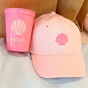 Peut inclure: Un sweat-shirt bleu clair avec le mot "SEASIDE" imprimé en bleu clair. Une bouteille d'eau rose avec le nom "SARAH" imprimé dessus. Un gobelet rose avec un motif de coquillage et le nom "SARAH" imprimé dessus. Une casquette de baseball rose avec un motif de coquillage.