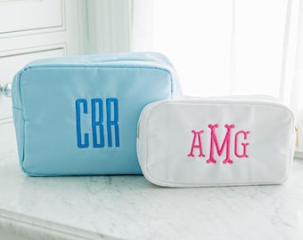 May include: A light blue rectangular pouch with the blue embroidered monogram CBR and a smaller white rectangular pouch with the pink embroidered monogram AMG. Both pouches feature a zipper closure and a structured, smooth fabric exterior.