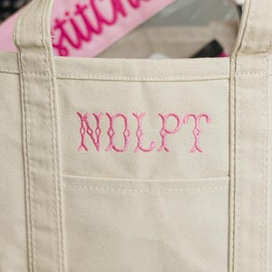 May include: A beige canvas tote bag with a pink embroidered monogram and a pink zippered pouch inside. The pouch has the word "stitches" embroidered in pink script. The tote bag has a front pocket.