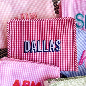 May include: A collection of rectangular zippered pouches featuring a gingham pattern in various colors including red, light pink, blue, and green. Each pouch is personalized with embroidered text such as DALLAS, AGM, and other initials in bold fonts.