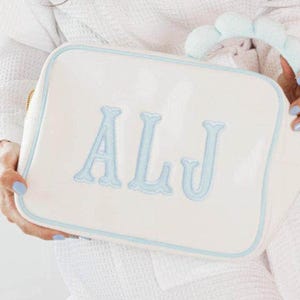 May include: A rectangular white cosmetic bag with rounded corners and light blue piping. The front features the embroidered light blue letters ALJ in a serif font. A matching light blue plush handle is attached to the top of the bag.