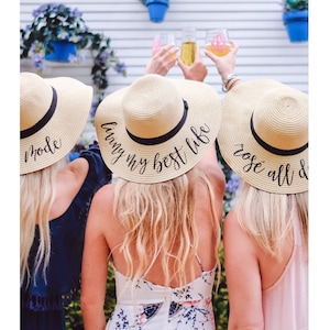 May include: Three friends wearing straw hats with different sayings on them. The hats say "living my best life", "rose all day", and "mode".