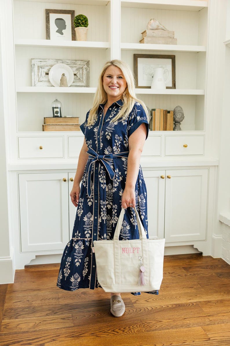 May include: A navy blue dress with a white floral pattern and a matching belt. The dress has short sleeves and a button-down front. A cream-colored tote bag with the letters "NOLPT" is held in front of the dress. The bag has a pink tassel.