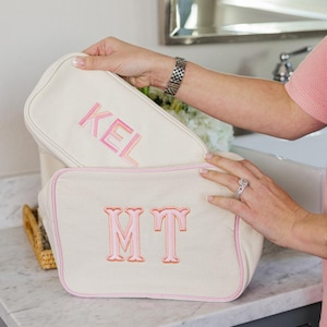 Könnte beinhalten: Zwei cremeweiße Stofftaschen mit rosafarbenen Paspeln. Die obere Tasche zeigt den gestickten rosafarbenen Schriftzug KEL, während die untere Tasche das gestickte rosafarbene Monogramm MT aufweist. Beide rechteckigen Taschen haben einen Reißverschluss.