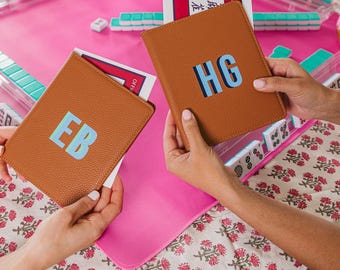 May include: Two tan, textured leather-like card holders featuring personalized monograms. One holder displays the letters EB in light blue and green, while the other shows HG in light blue and dark blue. Each holder contains a white card with text.