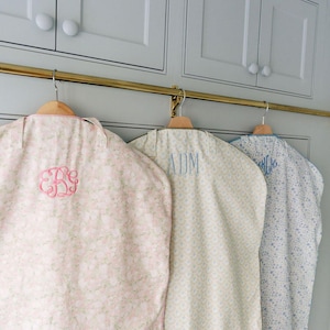 May include: Three garment bags hanging on a brass rod. The bags are patterned with floral and geometric designs in pastel colors. Each bag has a monogram in a contrasting color.
