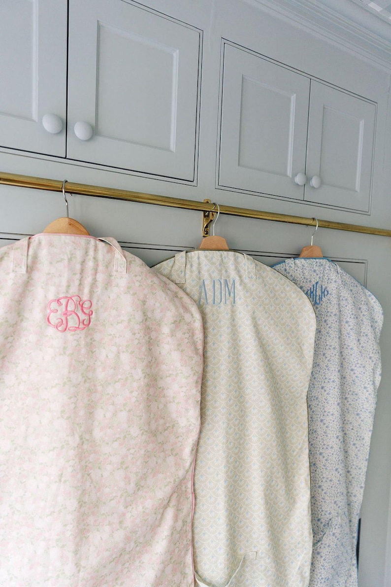 May include: Three garment bags hanging on a brass rod. The bags are patterned with floral and geometric designs in pastel colors. Each bag has a monogram in a contrasting color.