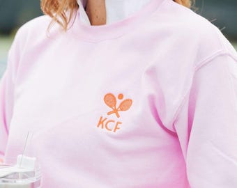 May include: A pink sweatshirt with a white collar and an orange embroidered logo of two tennis rackets and a ball with the letters "KCF" underneath.