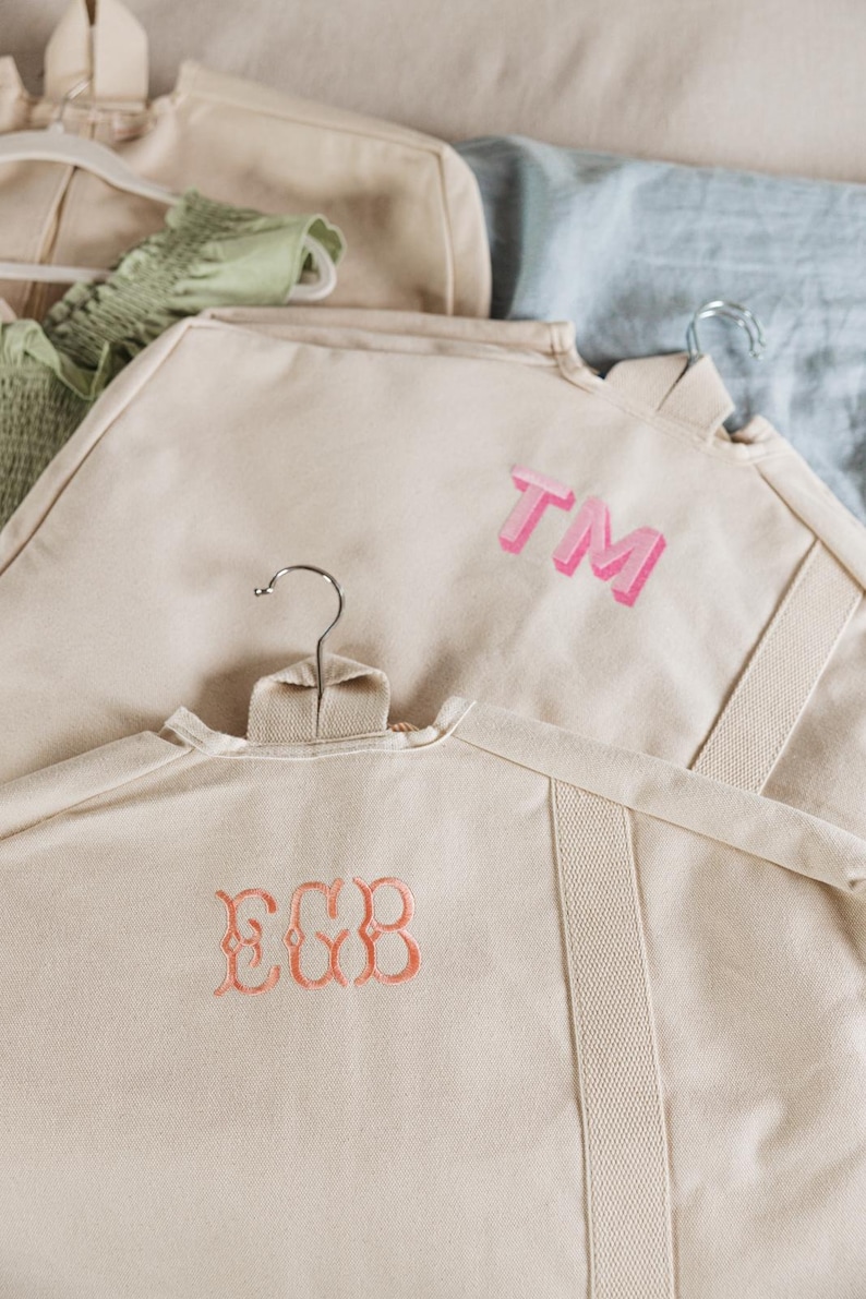 May include: Two garment bags with embroidered initials. The top bag has the initials 'TM' in pink. The bottom bag has the initials 'EGB' in orange. Both bags are made of a light beige canvas material.