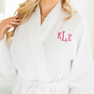 May include: White waffle-weave robe with a matching belt. The robe features a monogram in pink letters on the chest. The robe is designed with a shawl collar and pockets.